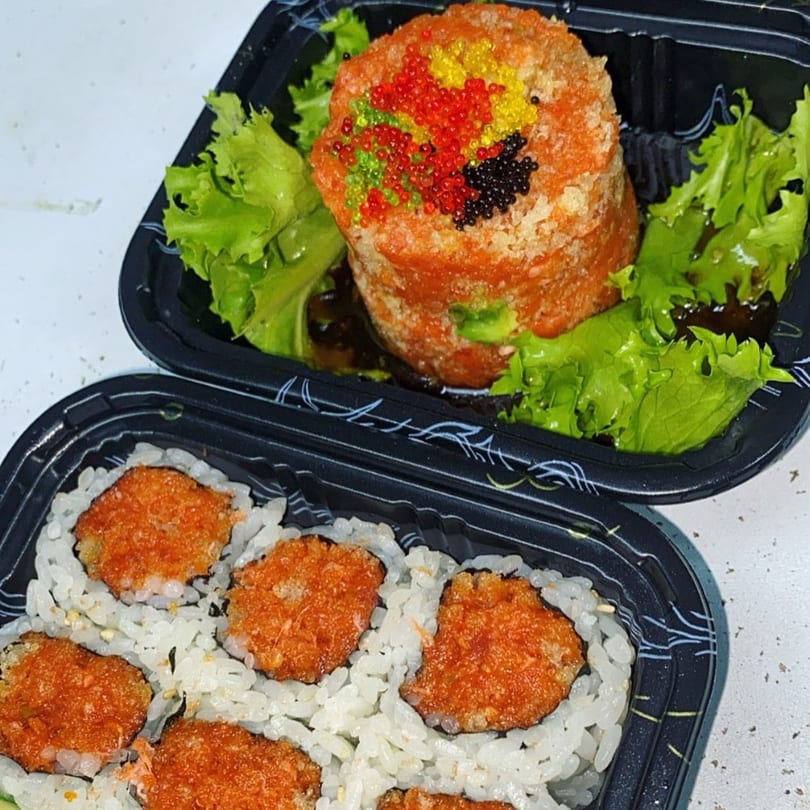 Two black takeout containers of Saga Fusion sushi, one with a tall roll topped with colorful roe.